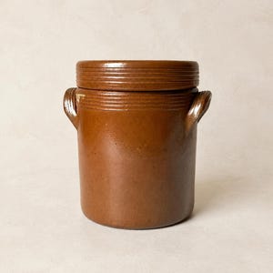 Può includere: Un vaso cilindrico in gres marrone con coperchio abbinato e due piccole maniglie laterali. La superficie esterna presenta una finitura liscia e smaltata con sottili dettagli a coste orizzontali lungo il bordo superiore e il coperchio.