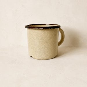 Könnte beinhalten: Ein gesprenkelter beiger Emaillebecher mit dunkelbraunem Rand und passendem Henkel. Der zylindrische Becher hat eine glatte Oberfläche mit einem kleinen dunklen Fleck nahe der Basis und zeigt ein klassisches Design mit robuster Konstruktion.