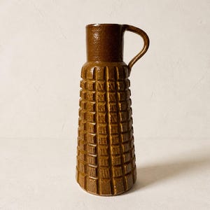 Może przedstawiać: Wysoki, cylindryczny brązowy dzbanek ceramiczny z błyszczącym wykończeniem. Korpus posiada powtarzający się wzór siatki z wgłębionymi kwadratami. Posiada gładką, niezdobioną szyjkę oraz pojedynczy zakrzywiony uchwyt przymocowany do górnej części.