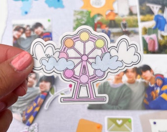 May include: A sticker of a pink and white Ferris wheel with rainbow colored gondolas. The Ferris wheel is surrounded by white clouds with blue accents. The sticker is on a white background.