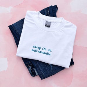 Peut inclure: Un t-shirt blanc à manches courtes avec un col rond, orné du texte brodé "sorry i'm an anti-romantic" en fil bleu sarcelle foncé. Le t-shirt est plié et superposé sur un jean en denim bleu foncé.
