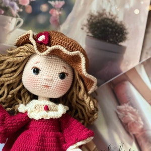 May include: A crocheted doll wearing a red dress, brown hat with a white trim, and a white lace collar. The doll has brown hair and blue eyes. The doll is sitting on a surface with a blurred background.