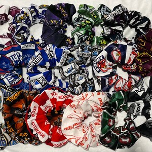 May include: A collection of 17 colourful hair scrunchies featuring various sports team logos. The scrunchies are made of fabric and have a gathered, ruffled look. The logos include the names of teams such as "Tigers", "Bulldogs", "St. George", and "Warriors".