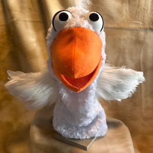 May include: White plush hand puppet with large black eyes and an orange beak. The puppet is designed to look like a bird.