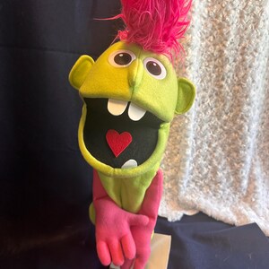 May include: A green hand puppet with a pink mohawk, large eyes, and a wide open mouth. The puppet is wearing a pink glove and has a red heart in its mouth.