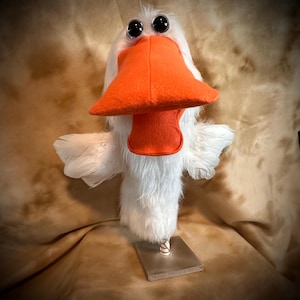May include: A white hand puppet with a large orange beak and black eyes. The puppet is standing on a wooden base.