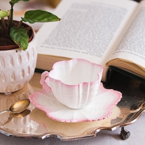 May include: A white teacup and saucer with a pink scalloped edge sit on a silver tray, next to a small potted plant and an open book. The teacup has a matching pink rim and handle. A gold spoon rests on the tray.