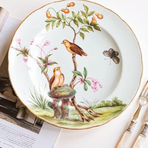May include: A round white ceramic plate with a scalloped gold rim, featuring a hand-painted scene of two brown birds perched on a tree branch with orange fruit, pink blossoms, green grass, and a single butterfly on the right side of the plate.