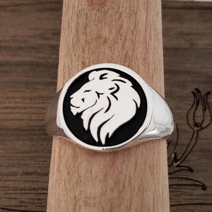 Op de afbeelding: Een zilveren zegelring met een witte leeuwenkop op een zwarte achtergrond. De ring heeft een gepolijste afwerking en een afgeronde vorm. De leeuwenkop is een gedetailleerd, gestileerd ontwerp.