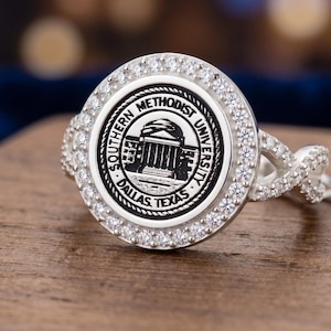 Puede incluir: Un anillo de plata con una cara circular que presenta el sello de la Southern Methodist University, incluyendo el texto Southern Methodist University Dallas Texas. El centro está rodeado por un halo de pequeñas piedras claras.