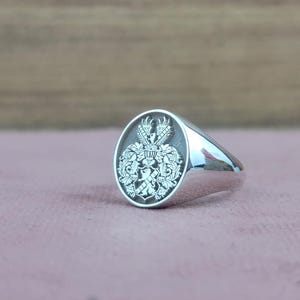 Op de afbeelding: Een zilveren zegelring met een gedetailleerd wapenontwerp. De ring heeft een gepolijste band en een ovale face met een gegraveerd heraldisch embleem. Het wapen bevat ingewikkelde details van een kroon, vleugels en andere sierlijke elementen.