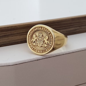 May include: A gold ring with a round, raised design featuring the Pennsylvania State University seal and the text "The Pennsylvania State University 1855".