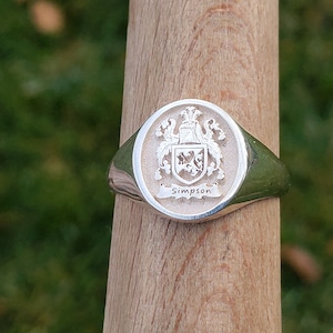 May include: Silver signet ring with an oval face. The ring features a detailed family crest with the name "Simpson" engraved below. The ring is displayed on a wooden dowel against a blurred green background.