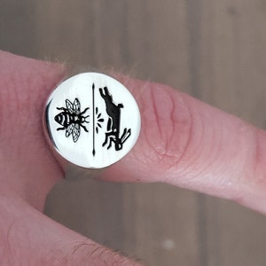 Op de afbeelding: Een zilveren ring met een ronde voorkant met een zwarte bij en een zwarte illustratie van een konijn en een plant. De ring wordt op een vinger getoond.