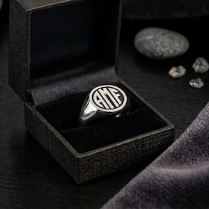 May include: A silver signet ring featuring a circular face engraved with the letters AMF in a bold, block-style monogram. The ring is displayed inside a black, textured, square-shaped jewelry box with a hinged lid and a black velvet interior.