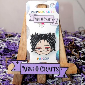 May include: A purple and black banner with the text "Mini Q Crafts" on a wooden easel. The banner is decorated with small diamonds. A black and white cartoon character with red eyes is on a PopSocket.