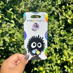 May include: A black PopSocket with a cartoon-like character featuring large white eyes and colorful flower accents. The PopSocket is attached to a white card with the "POPSOCKETS" logo and a small graphic.