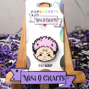May include: A pink and black PopSocket featuring a cartoon character with pink hair and a serious expression. The PopSocket is packaged in a white and silver card with the text "PopSockets Mini Q Crafts" printed on it.