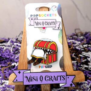 May include: A purple and white banner with the text "Mini Q Crafts" on a wooden easel. The banner is in front of a white and silver PopSocket with a cartoon image of a person reaching into a treasure chest.