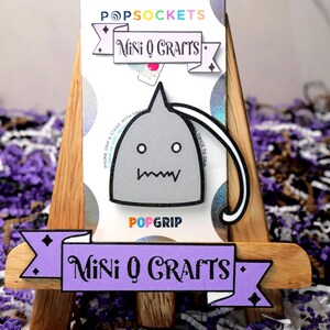 May include: A gray and white PopSocket phone grip with a cartoon monster design. The PopSocket is on a wooden easel with a purple banner that reads "Mini Q Crafts".
