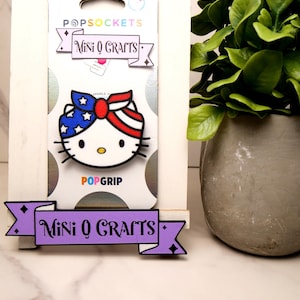 May include: A purple banner with the text "Mini Q Crafts" and black diamond shapes on either end. The banner is in front of a white background. A white and red PopSocket with a Hello Kitty design wearing a red, white, and blue bandana is on a white card with the text "PopSockets" and "PopGrip".