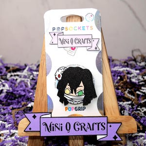 May include: A black and green popsocket featuring a cartoon character with a bandage over their eyes. The popsocket is on a wooden easel with a purple banner that says "Mini Q Crafts".