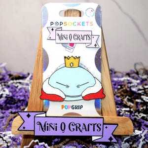 May include: A PopSockets PopGrip featuring a light blue cartoon character wearing a gold crown and red and white robes. The PopGrip is on a white backing with the text "Mini O Crafts" and "PopSockets". The PopGrip is displayed on a wooden stand.