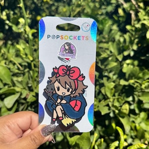 May include: A PopSockets phone grip featuring a cartoon character with a red bow, holding a black cat on a broom. The grip is on a white and iridescent backing with the PopSockets logo and a small sticker.