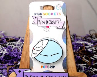 May include: A blue and white PopSocket with a cartoon face and the text "Mini Q Crafts" on a purple banner. The PopSocket is on a wooden easel.