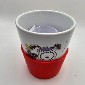 May include: White ceramic mug with a black and white cartoon dog wearing a purple beret. The mug has a red silicone sleeve.