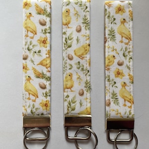 May include: Three fabric keychains with a spring-themed pattern. The white fabric is decorated with yellow ducklings, eggs, and green foliage. Each keychain has a gold-colored metal clasp and a silver-colored key ring.