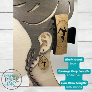 May include: A wooden display featuring a gymnastics-themed hair claw and earrings. The hair claw is 3.35 inches long and the earrings have a 2.5-inch drop. Both items are engraved with the word "Gymnastics" and a gymnast silhouette.