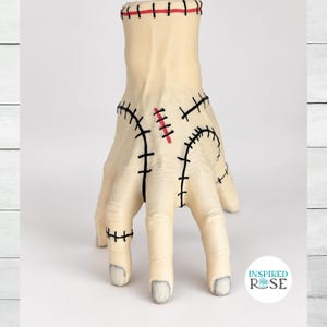 May include: A pale, beige-colored hand sculpture featuring prominent black stitched markings across the fingers, palm, and wrist. A single red stitch is visible on the back of the hand, and the fingernails are painted a muted gray color.