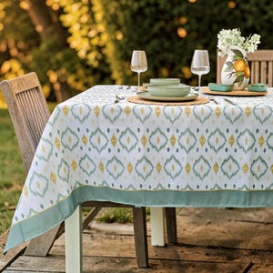 Pode incluir: Uma toalha de mesa branca com um padrão repetido de diamantes amarelos e verde-azuis cobre uma mesa de madeira montada para jantar. A mesa está montada com pratos, copos e um vaso de flores. Uma borda verde-azulado adorna a toalha de mesa.