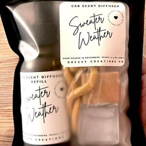 May include: A clear glass bottle with a white label that says "Car Scent Diffuser Refill Sweater Weather" and a small heart icon. The bottle is in a clear plastic bag with a brown wooden car air freshener and a white label that says "Car Scent Diffuser Sweater Weather" and a small heart icon. The label also says "Hand Poured in Rosenberg, Texas | 1.5 oz (44ml) Dressy Creations Co."