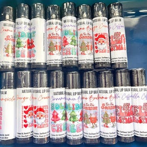 May include: A collection of 12 natural lip balms with various holiday-themed designs. The lip balms are black with white labels and feature designs such as "Tis the Season", "Santa", "Jingle Bells", and "Holiday Cheer".