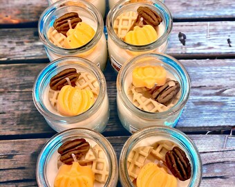 May include: A collection of small glass jars filled with a white candle and topped with fall-themed decorations. The decorations include small pumpkin shapes, waffle pieces, and pecan halves.