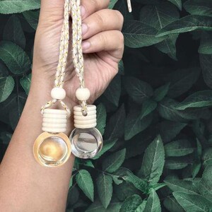 May include: Two small, round glass bottles with wooden tops and hanging cords. One bottle contains a golden liquid, the other is clear. The bottles are suspended against a backdrop of green leaves, suggesting a natural or aromatherapy theme.