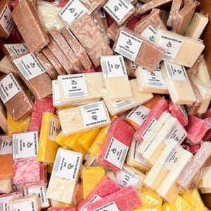 May include: A variety of artisan cold process soaps in various colors and scents. The soaps are wrapped in clear plastic and labeled with the scent and ingredients. The soaps are arranged in a pile, with some of the soaps overlapping.