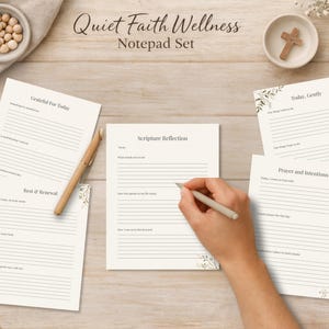 May include: A notepad set with the text "Quiet Faith Wellness" and "Notepad Set". The set includes several lined pages with prompts for reflection, gratitude, and prayer. A hand is holding a pen over one of the pages. The pages have floral accents.