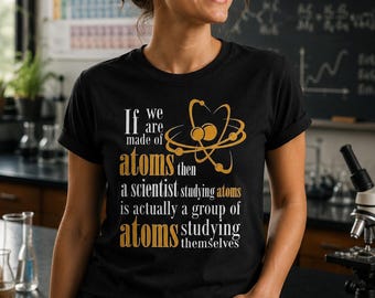 Puede incluir: Una camiseta negra de manga corta con un gráfico dorado de un átomo y el texto: If we are made of atoms then a scientist studying atoms is actually a group of atoms studying themselves. La camiseta tiene cuello redondo y mangas remangadas.