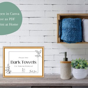 Puede incluir: Un cartel enmarcado con fondo blanco y texto negro que dice "Por favor, use toallas oscuras para quitarse el maquillaje Gracias". El cartel est&aacute; en un estante de madera en un ba&ntilde;o con paredes de azulejos blancos. Hay toallas, un dispensador de jab&oacute;n y una planta en maceta en el estante.