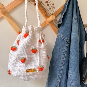May include: White crocheted drawstring bag with red and green strawberry embellishments. The bag has a gold label with the text "Pretty Art".