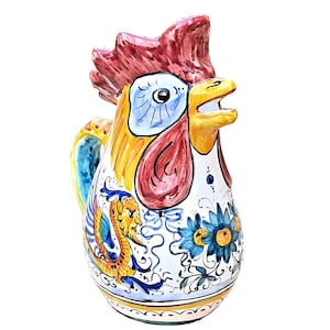 May include: A ceramic rooster pitcher with a yellow, red, blue and white painted design. The pitcher features a rooster head with a large beak and a floral pattern.