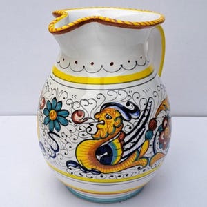 May include: White ceramic jug with a yellow handle and rim. The jug is decorated with a colourful design featuring a mythical creature, flowers, and swirling patterns. The base has a blue and yellow stripe.