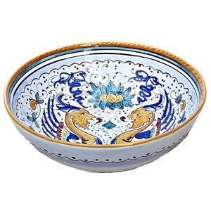 May include: A white ceramic bowl with a blue and gold floral design. The bowl features two stylized animal heads with wings and a central floral motif.