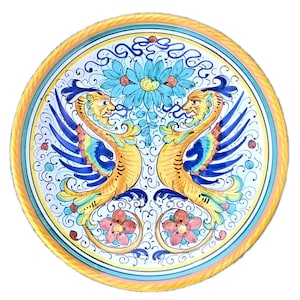 May include: A round ceramic plate with a yellow border and a white centre. The centre features a blue and yellow design of two winged creatures with a floral motif in the centre.