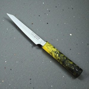 May include: A stainless steel paring knife with a yellow and black marbled handle. The knife blade is silver and has the text "KOCAKAYA - Life Series" printed on it. The knife is angled on a gray surface.