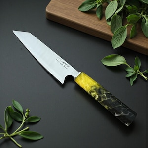 May include: A stainless steel chef's knife with a yellow and black marbled handle. The blade is angled and has the text "KOCAKAYA Life Series" etched on it. The knife is on a black surface with green herbs and a wooden cutting board in the background.