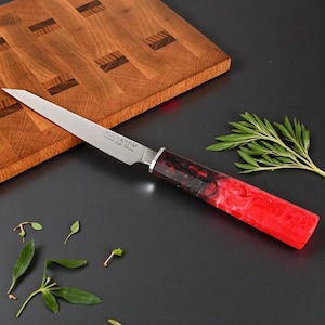 Könnte beinhalten: Ein Edelstahlmesser mit einem rot-schwarz marmorierten Griff. Die Messerklinge ist abgewinkelt und scharf, mit dem Markennamen "KOCAKAYA - Life Series" auf der Klinge. Das Messer liegt auf einer dunklen Oberfläche mit einem Holzschneidebrett im Hintergrund.